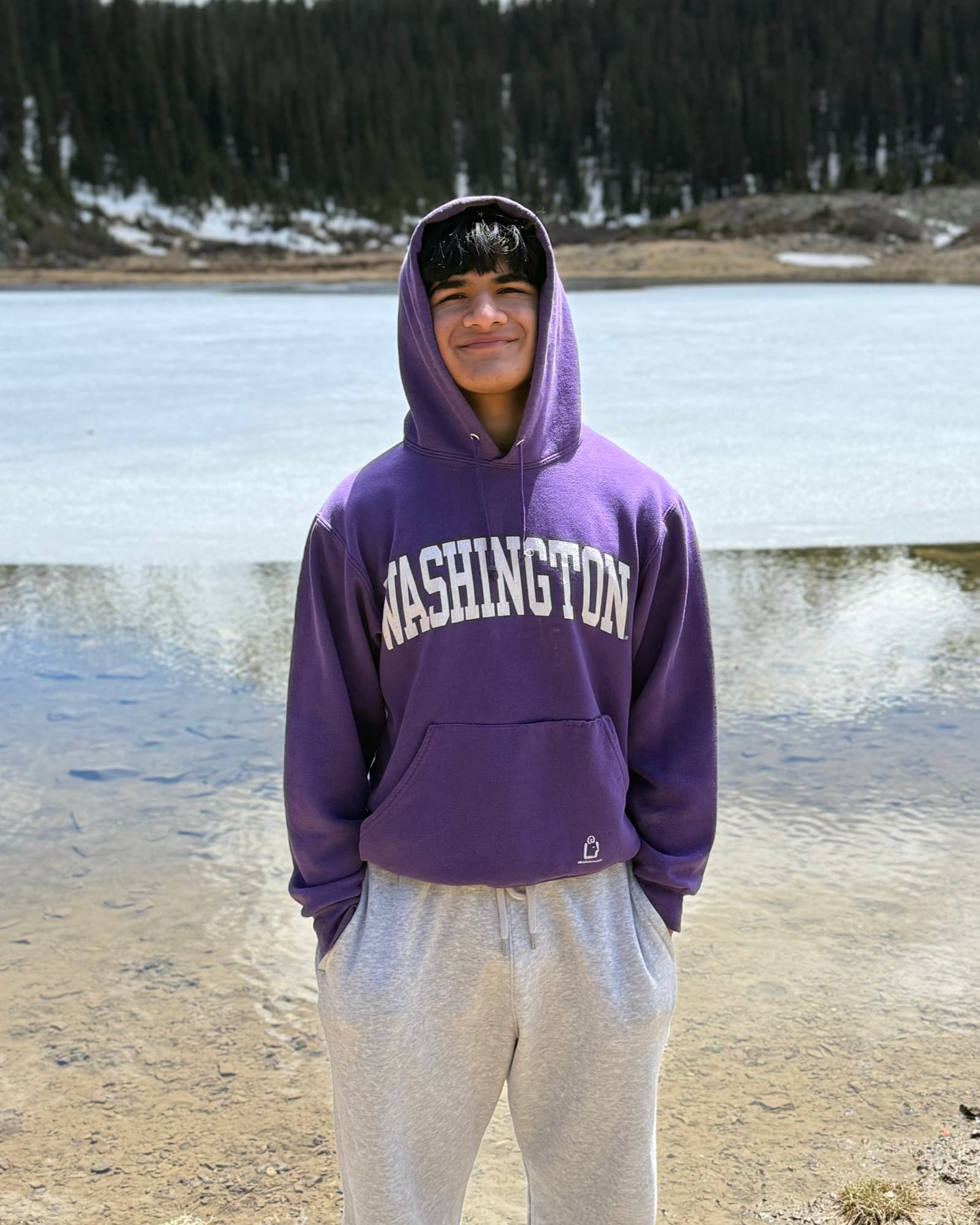 Aditya Kothuri standing in front of a partly frozen alpine lake, with a pine forest and a snow-covered mountain ridge behind.