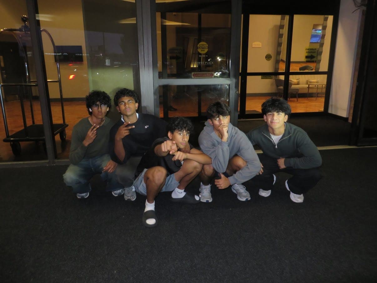 Five friends crouched shoulder to shoulder in a lit hotel doorway at night, all posing for the camera.
