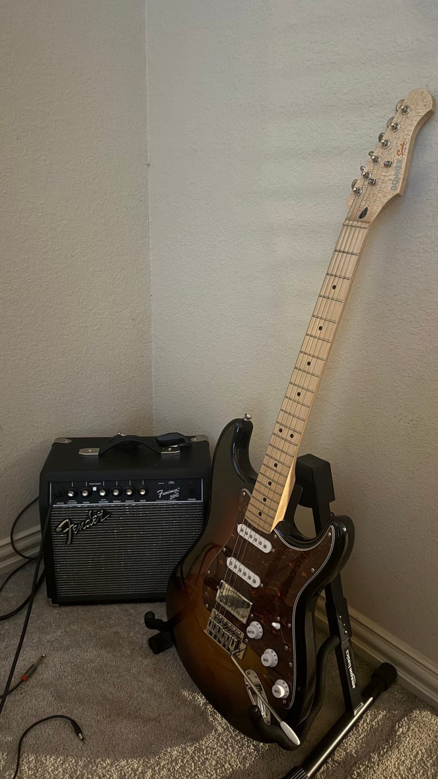 A sunburst electric guitar on a stand in the corner of a room, next to a small Fender practice amp.