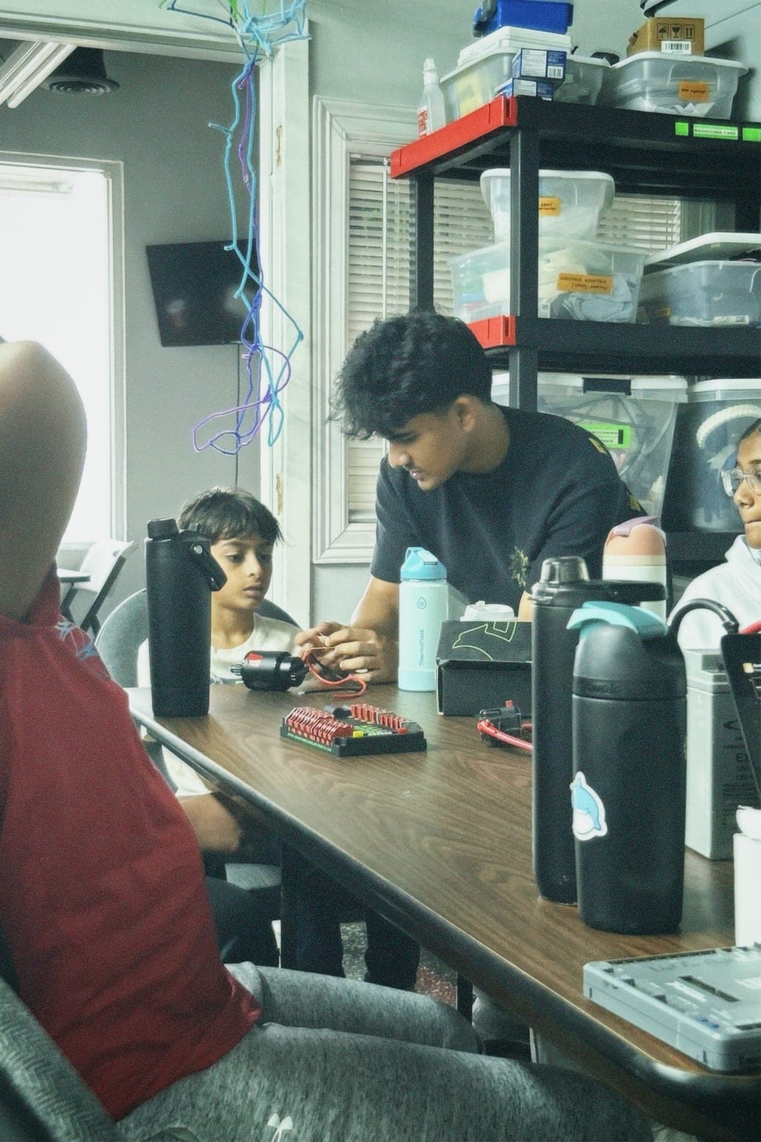 At a Radicubs summer camp workbench, a team member in a black T-shirt leans in beside a younger camper over a small robot's electronics, parts bins shelved behind them.