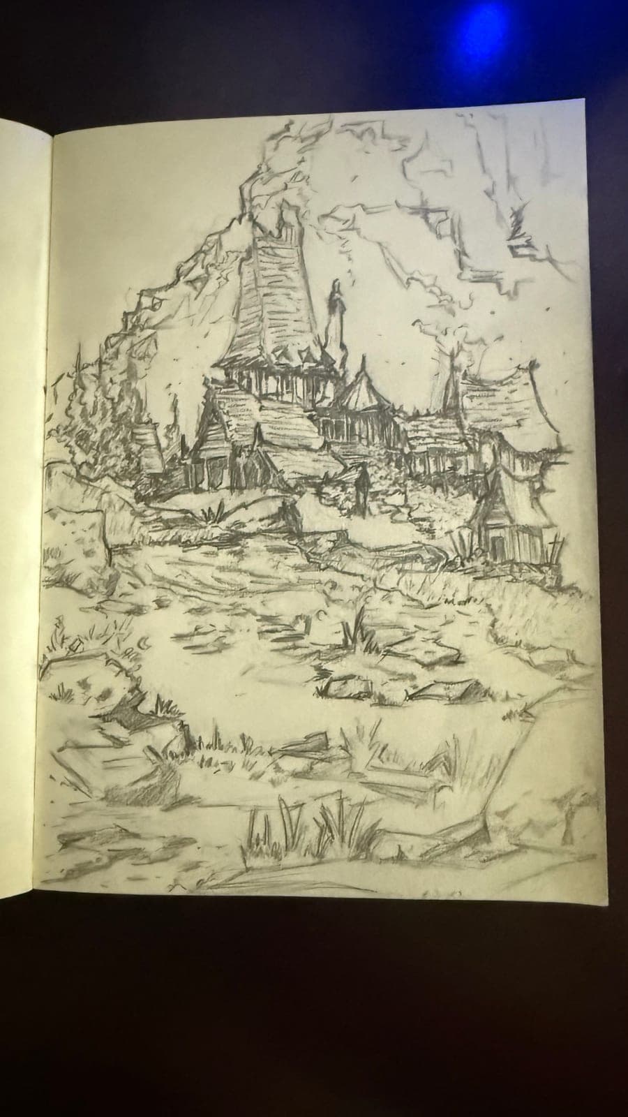 Pencil sketch in a sketchbook: a cluster of steep-roofed houses and a tower built against a cliff, with a stony path leading up to them.